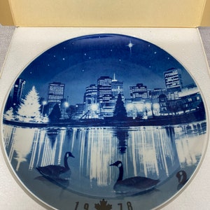 May include: A blue and white ceramic plate depicting a cityscape reflected in water. The plate features two geese swimming in the water and the text "1978" with a Canadian maple leaf.