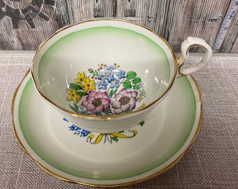 May include: A vintage teacup and saucer set with a floral design. The cup and saucer are white with a green rim and a gold trim. The floral design features pink, yellow, and blue flowers.