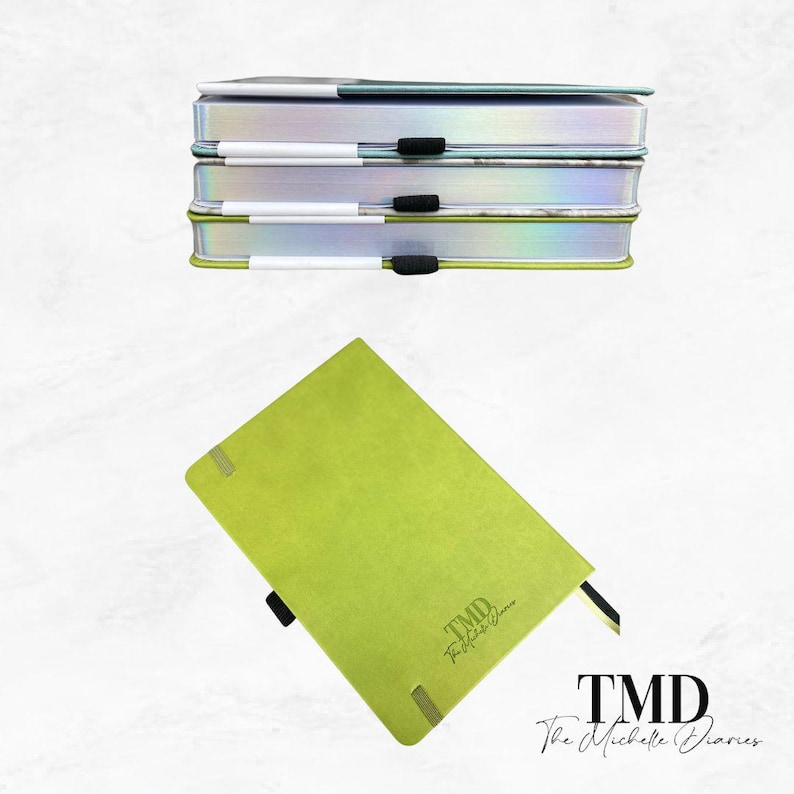 Op de afbeelding: Een stapel van drie groene dagboeken met de tekst "TMD The Michelle Diaries" op de voorkant. De dagboeken hebben een holografische cover en een zilveren pen.