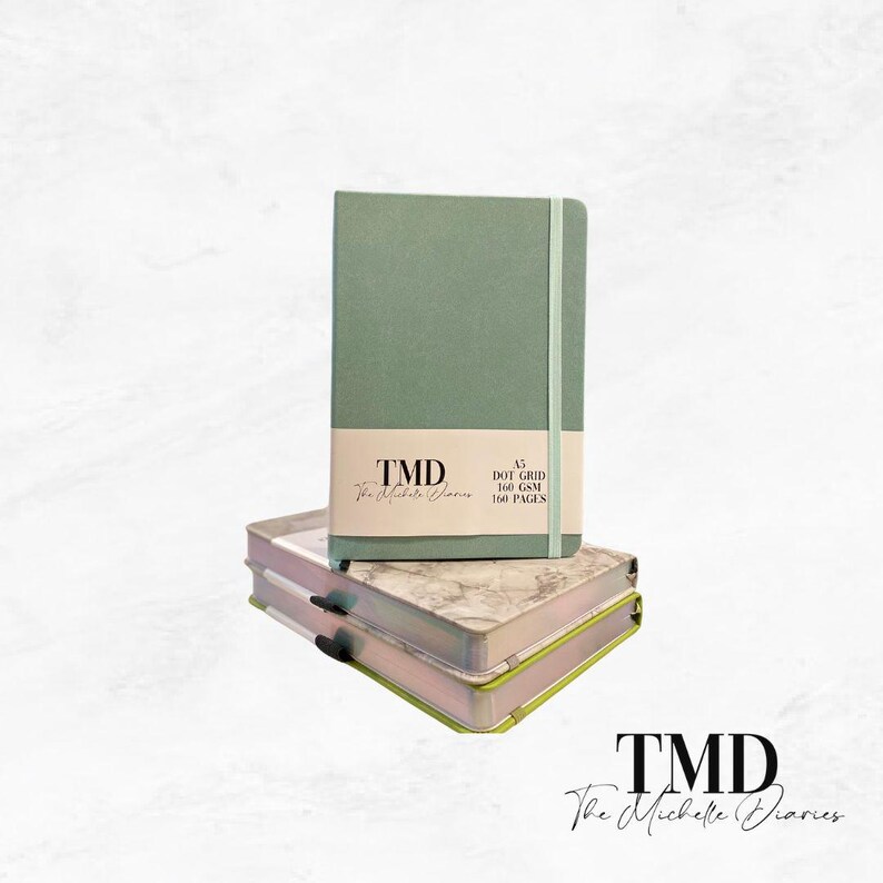 Op de afbeelding: Een groen A5-puntraster notitieboek met de tekst "TMD The Michelle Diaries" en "A5 DOT GRID 160 GSM 160 PAGES" gedrukt op de voorkant.
