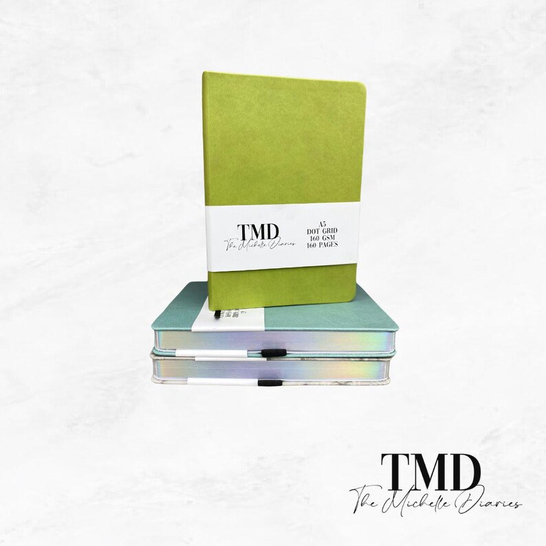 Op de afbeelding: Een stapel van drie notitieboekjes. Het bovenste notitieboekje is groen en heeft de tekst "TMD The Michelle Diaries A5 DOT GRID 160 GSM 100 PAGES" op de voorkant. De onderste twee notitieboekjes zijn blauw en hebben een holografische afwerking.
