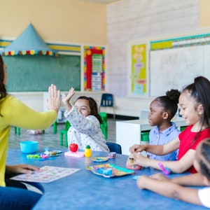 May include: A group of children are sitting at a table in a classroom. They are playing with toys and having fun. The teacher is interacting with the children by giving a high five.