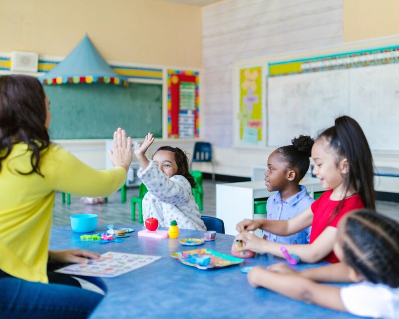 May include: A group of children are sitting at a table in a classroom. They are playing with toys and having fun. The teacher is interacting with the children by giving a high five.