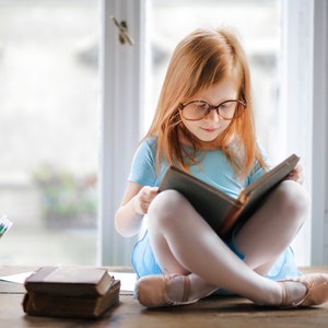Pu&ograve; includere: Una ragazza con i capelli rossi &egrave; seduta su una superficie di legno, indossa un vestito blu chiaro e occhiali, leggendo un libro.