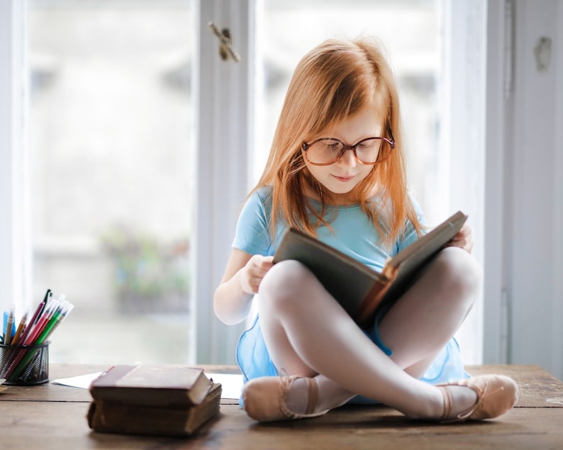 Pu&ograve; includere: Una ragazza con i capelli rossi &egrave; seduta su una superficie di legno, indossa un vestito blu chiaro e occhiali, leggendo un libro.