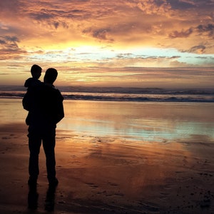 May include: Silhouette of a parent holding a child on their shoulders, standing on a sandy beach at sunset. The sky is a vibrant mix of orange, pink, and purple.