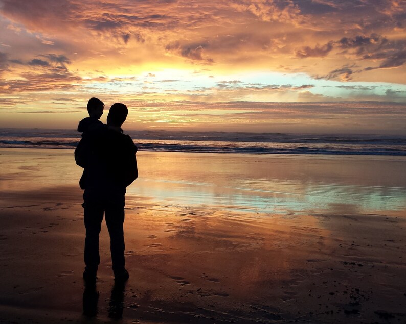 May include: Silhouette of a parent holding a child on their shoulders, standing on a sandy beach at sunset. The sky is a vibrant mix of orange, pink, and purple.