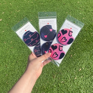 May include: Three sets of black and pink car coasters with heart designs. Each set is packaged in a clear plastic bag with a white label that reads "Car Coasters Handmade with love".
