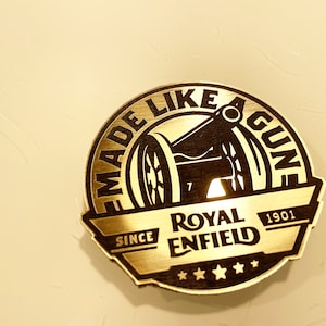 Könnte beinhalten: Goldenes Metallemblem mit einem schwarzen Kanonen-Design und dem Text "Made Like A Gun" über den Worten "Royal Enfield" und "Since 1901".