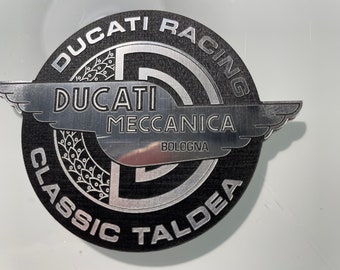 Op de afbeelding: Een rond, zwart en zilver Ducati Racing embleem. Het embleem bevat de woorden "DUCATI RACING", "DUCATI MECCANICA BOLOGNA" en "CLASSIC TALDEA" in zilveren letters. Het embleem heeft een getextureerde achtergrond en een vleugelontwerp.
