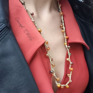 Op de afbeelding: Een close-up van een vrouw die een rood overhemd met kraag draagt met een gehäkelde ketting met bruine, oranje en gouden kralen. De ketting is gelaagd met een dunne gouden ketting. De tekst "Concept TOPlus" is zichtbaar op het overhemd.