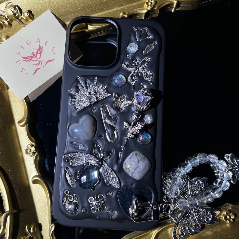 May include: Black phone case with a variety of silver and iridescent embellishments, including a crown, a heart, a butterfly, and a rose. The case has a black background and a clear, protective layer over the embellishments.