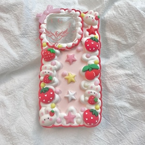 Op de afbeelding: Een roze en rode telefoonhoes met een wit en roze ontwerp. De hoes heeft een transparant venster voor het telefoonscherm en is versierd met aardbeien, sterren en konijntjes.
