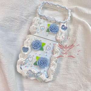 May include: A white phone case with a blue and white floral design. The case has a raised, 3D design that looks like whipped cream. There are also small, white pearls and glitter accents.