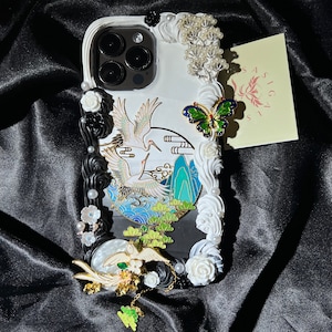 K&ouml;nnte beinhalten: Wei&szlig;es Handy-Case mit einem schwarz-wei&szlig;en Kranich-Design, wei&szlig;en Blumen und einem gr&uuml;nen Schmetterling. Das Case hat auch eine transparente R&uuml;ckseite mit einem Kameraobjektiv.