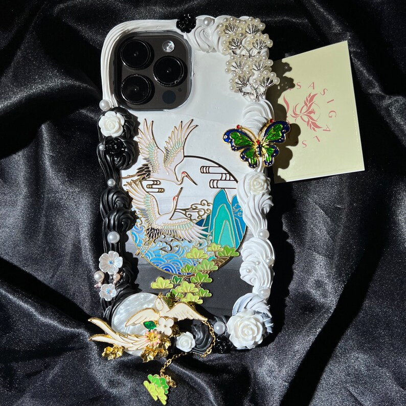 K&ouml;nnte beinhalten: Wei&szlig;es Handy-Case mit einem schwarz-wei&szlig;en Kranich-Design, wei&szlig;en Blumen und einem gr&uuml;nen Schmetterling. Das Case hat auch eine transparente R&uuml;ckseite mit einem Kameraobjektiv.