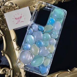 May include: A clear phone case with a design of blue, green, and white glitter embedded in resin. The glitter is shaped like small pebbles.