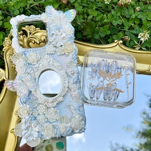 Puede incluir: Una funda para teléfono blanca y azul con un espejo y adornos florales. La funda está decorada con una mariposa y purpurina. También se muestra una funda de plástico transparente con detalles dorados.