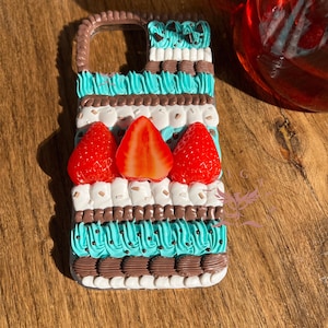 Könnte beinhalten: Eine Handyhülle, verziert mit texturiertem, aufgespritztem Zuckerguss in Schichten aus Braun, Weiß und hellem Türkis. Das Design zeigt drei realistische rote Erdbeerhälften, die horizontal in der Mitte platziert und mit braunen Streuseln verziert sind.