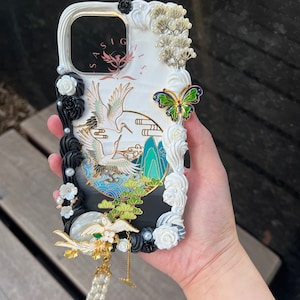 K&ouml;nnte beinhalten: Eine wei&szlig;e und schwarze Handyh&uuml;lle mit einem Kranich-Design, wei&szlig;en Blumen und einem gr&uuml;nen Schmetterlings-Anh&auml;nger. Die H&uuml;lle hat einen schwarzen Hintergrund mit einem wei&szlig;en Rand und einem wei&szlig;en Kranich-Design. Die H&uuml;lle hat auch einen wei&szlig;en Schmetterlings-Anh&auml;nger mit gr&uuml;nen Fl&uuml;geln.