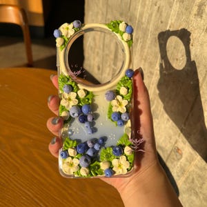 Puede incluir: Funda transparente para teléfono decorada con arándanos, flores blancas y hojas verdes. La funda tiene un recorte circular para la cámara y está adornada con pequeñas cuentas transparentes. El diseño es tridimensional y detallado.