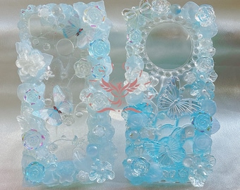 May include: Two clear blue resin phone cases with a floral and butterfly design. The cases are decorated with 3D butterflies, flowers, and pearls.