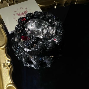 Puede incluir: Un objeto decorativo gris oscuro detallado con una pieza central de calavera, acentuado con piedras preciosas rojas y transparentes. El objeto tiene diseños intrincados y arremolinados y está sobre una superficie negra. Una tarjeta con el texto "SASIGALS" es visible.
