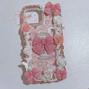 May include: A pink and white phone case with a floral design. The case features a pink bow, white flowers, and pearl accents.