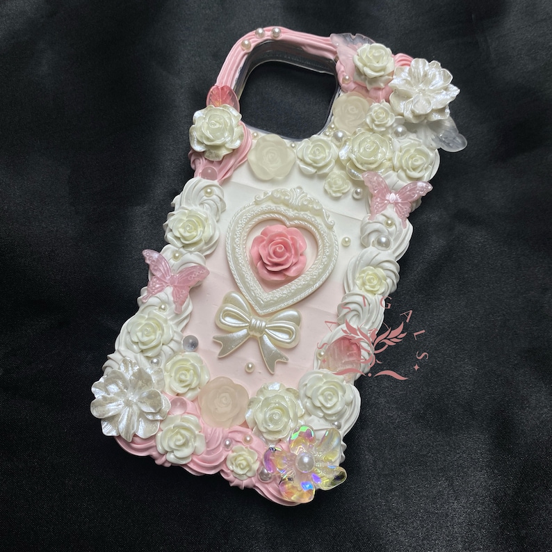 Op de afbeelding: Een roze en witte telefoonhoesje versierd met 3D-bloemen, parels en een roze hart met een strik. De hoes heeft een transparante achterkant om de telefoon te laten zien.