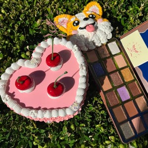 Puede incluir: Un pastel rosa en forma de corazón con glaseado blanco y tres cerezas rojas en la parte superior. Una paleta de sombras de ojos dorada y verde con un diseño floral está al lado del pastel.