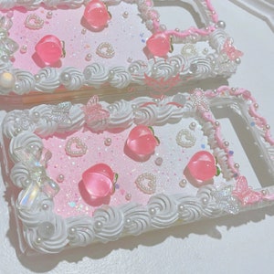 Puede incluir: Dos fundas de teléfono rosa y blancas decoradas con cuentas de perlas, dijes de melocotón, purpurina y mariposas.