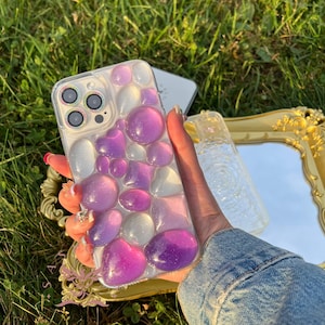 Op de afbeelding: Een transparante telefoonhoes met een ontwerp van paarse en witte harsachtige vormen. De hoes is ontworpen voor een telefoon met een drievoudige lenscamera.