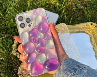 Op de afbeelding: Een transparante telefoonhoes met een ontwerp van paarse en witte harsachtige vormen. De hoes is ontworpen voor een telefoon met een drievoudige lenscamera.