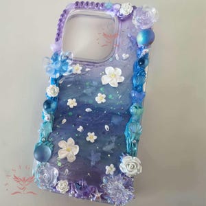 Könnte beinhalten: Dekorative Handyhülle mit blau-violettem Farbverlauf, verziert mit weißen Blumen, blauen Muscheln und transparenten, funkelnden Verzierungen. Die Hülle hat einen transparenten Rand mit lila Perlen und floralen Akzenten.