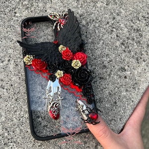 Peut inclure: Une coque de téléphone noire avec un motif floral noir et rouge, une main argentée tenant deux épées et un cœur rouge. La coque présente également une araignée noire avec un bijou rouge sur le dos.