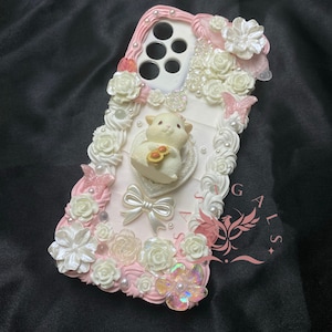 May include: A white and pink phone case decorated with white roses, pearls, and a small white lamb figurine. The case has a pink and white swirl design and the text "GALS" is written on the side.