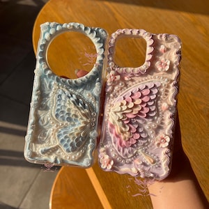 Peut inclure: Deux coques de téléphone avec des motifs en relief texturés. La coque de gauche est bleu clair avec un papillon et des perles. La coque de droite est rose et blanche avec un motif d'ailes de papillon, des petites fleurs et des perles.