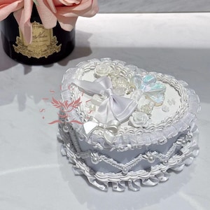 Puede incluir: Caja de corazón blanca con un borde de encaje blanco y un lazo blanco. La caja está decorada con flores blancas y una mariposa iridiscente.