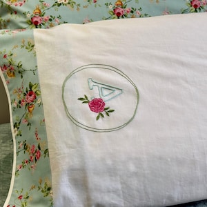 May include: White pillowcase with a green embroidered circle containing the letter "A" and a pink flower. The pillowcase has a floral border with pink and white flowers on a light blue background.