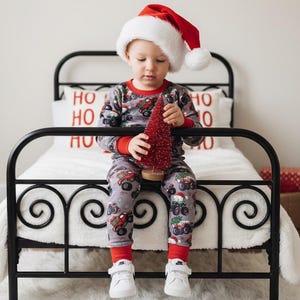 May include: A child wearing a Santa hat and Christmas pajamas sits on a black metal bed frame. The pajamas are gray with red accents and feature a tractor design. The child holds a small red Christmas tree.