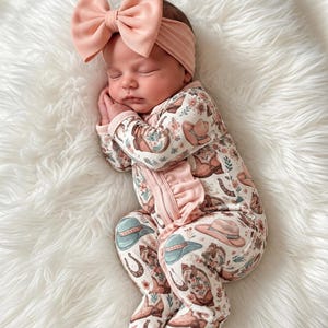 May include: A long-sleeved, footed baby sleepsuit with a ruffled front zip fastening, featuring a repeating pattern of cowboy hats, boots, horseshoes, and flowers. The sleepsuit is paired with a matching solid peach-coloured headband with a large bow. Image created with AI.