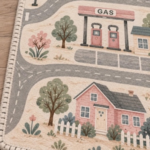 May include: A rectangular area rug featuring a printed town scene with gray roads, a pink house with a white picket fence, a gas station with the text GAS, green trees, pink flowering bushes, a traffic light, and a small boat on blue water.
