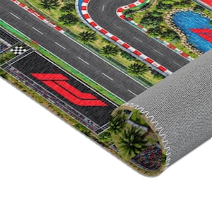May include: A rolled floor mat featuring a printed graphic of a Formula 1 style racetrack with gray asphalt, red and white curbing, green landscaping, a blue water feature, and a large red logo on the track surface, shown from a high angle.