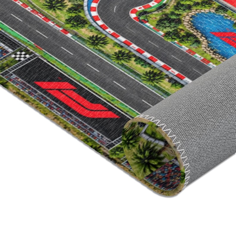 May include: A rolled floor mat featuring a printed graphic of a Formula 1 style racetrack with gray asphalt, red and white curbing, green landscaping, a blue water feature, and a large red logo on the track surface, shown from a high angle.