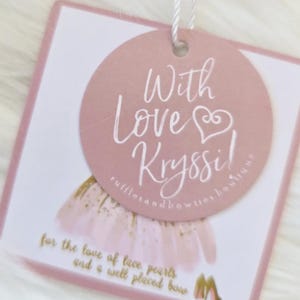 May include: A pink and white tag with the text "With Love Kryssi!" in cursive script. The tag also includes the text "rufflesandbowties boutique" and "for the love of lace pearls and a well placed bow."