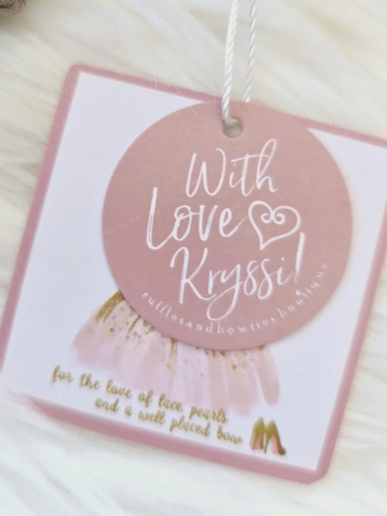 May include: A pink and white tag with the text "With Love Kryssi!" in cursive script. The tag also includes the text "rufflesandbowties boutique" and "for the love of lace pearls and a well placed bow."