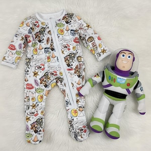 May include: A white long-sleeved footed sleeper featuring a colorful repeating pattern of Toy Story characters and icons, including Buzz Lightyear, Woody, and green aliens, with a full-length front zipper, alongside a Buzz Lightyear action figure.