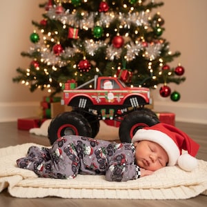 May include: A sleeping baby wearing a gray onesie and a Santa hat, lying on a cream-colored blanket. A red monster truck with Christmas decorations sits behind the baby, in front of a decorated Christmas tree.