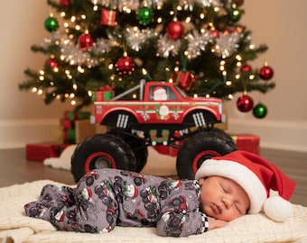 May include: A sleeping baby wearing a gray onesie and a Santa hat, lying on a cream-colored blanket. A red monster truck with Christmas decorations sits behind the baby, in front of a decorated Christmas tree.