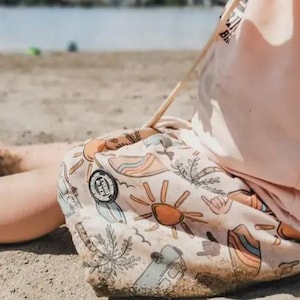 May include: A light pink beach bag featuring a pattern of suns, rainbows, surfboards, and palm trees. The bag has a round patch with text and a drawstring closure. The bag is on a sandy beach.